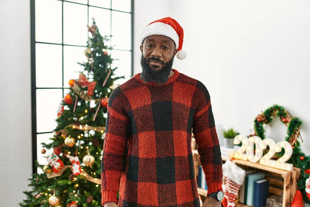 Αφροαμερικάνος που φοράει καπέλο Σάντα Κλάους και στέκεται δίπλα στο χριστουγεννιάτικο δέντρο χαλαρός με σοβαρή έκφραση στο πρόσωπο. απλή και φυσική κοιτάζοντας την κάμερα.  - Φωτογραφία, εικόνα