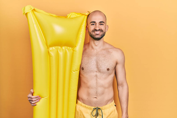 Mladý plešatý muž v plavkách a držení letní matrace float vypadá pozitivně a šťastně stojící a usmívá se s sebevědomým úsměvem ukazující zuby  - Fotografie, Obrázek