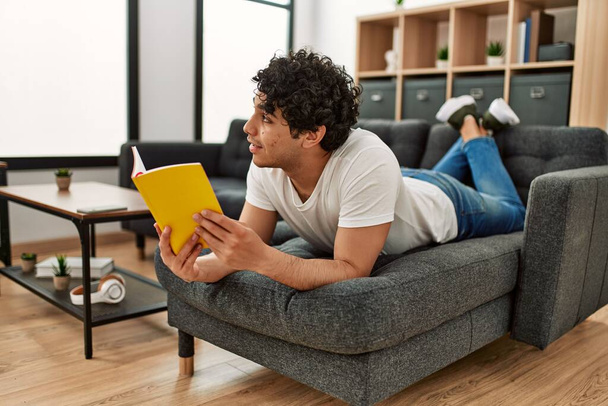 Νεαρός Ισπανός διαβάζει βιβλίο ξαπλωμένος στον καναπέ στο σπίτι. - Φωτογραφία, εικόνα