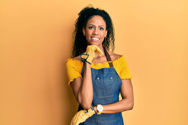 Middle age african american woman wearing professional apron with hand on chin thinking about question, pensive expression. smiling and thoughtful face. doubt concept.  - Foto, Imagen