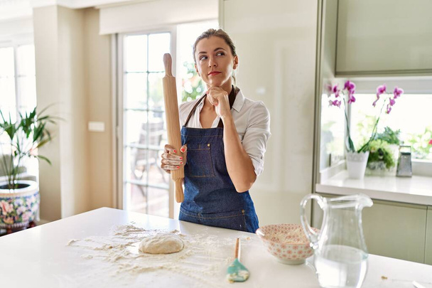 Belle femme blonde préparant la pâte pour la pizza visage sérieux en pensant à la question avec la main sur le menton, réfléchi sur l'idée déroutante  - Photo, image