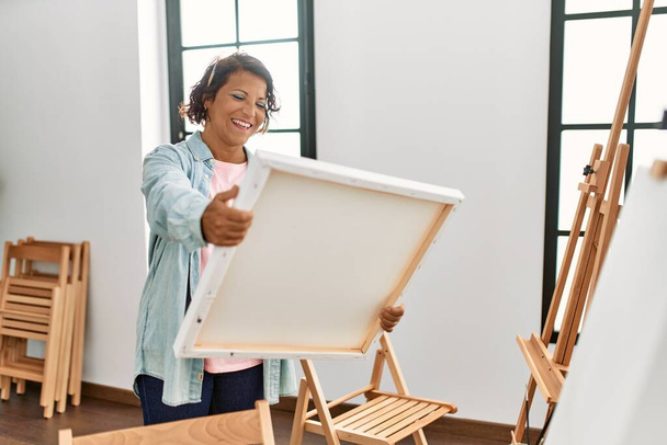 Middle age hispanic artist woman smiling happy holding canvas at art studio. - Фото, изображение
