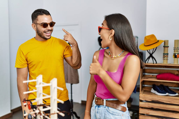 Νεαρό ζευγάρι Λατίνων χαμογελά χαρούμενος επιλέγοντας γυαλιά ηλίου στο κατάστημα ρούχων. - Φωτογραφία, εικόνα