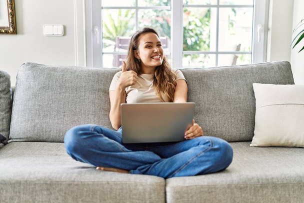 Güzel, genç, esmer bir kadın evdeki bilgisayarı kullanarak koltukta oturuyor ve elleriyle mutlu bir jest yapıyor. Kameraya bakıp başarıyı gösteren ifade.  - Fotoğraf, Görsel