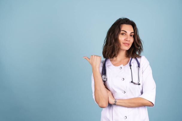 Giovane bella donna medico in un camice da laboratorio su uno sfondo blu sorride, e punta il dito a sinistra su uno spazio vuoto - Foto, immagini