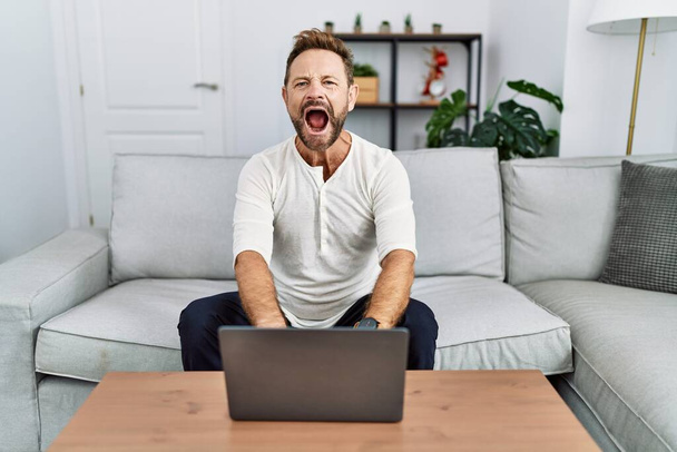 Mężczyzna w średnim wieku używający laptopa w domu wściekły i wściekły krzyczący sfrustrowany i wściekły, krzyczący ze złości. wściekłość i agresywne pojęcie.  - Zdjęcie, obraz