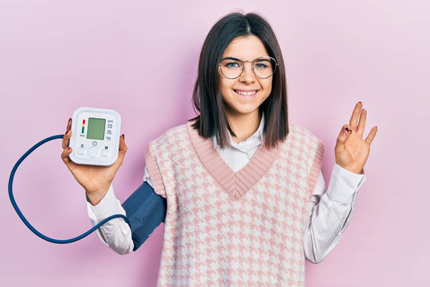 Mladá bruneta žena pomocí monitoru krevního tlaku dělá v pořádku znamení s prsty, s úsměvem přátelské gestikulace vynikající symbol  - Fotografie, Obrázek