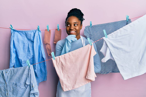 Africká Američanka s copánkovými vlasy praní oblečení na prádelní šňůře s rukou na bradě přemýšlet o otázce, zamyšlený výraz. usmívající se a zamyšlený obličej. pochybný koncept.  - Fotografie, Obrázek