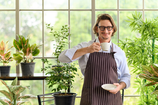 Portrait of happy senior man gardening wearing glasses and denim apron spending summer morning holding cup of hot coffee enjoying in the home gardening. looking at camera - Fotografie, Obrázek