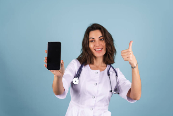 Junge schöne Ärztin in Labormantel auf blauem Hintergrund hält ein Telefon mit leerem schwarzen Bildschirm, lächelt, zeigt Daumen hoch - Foto, Bild