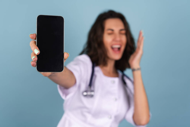 Junge schöne Ärztin in Labormantel auf blauem Hintergrund, ein Telefon mit leerem schwarzen Bildschirm in der Hand, überrascht schreiend, schockiert - Foto, Bild