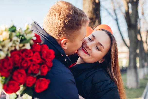 Valentines day date. Man kissing girlfriend on cheek outdoors gifting bouquet of red roses. Couple in love walking in city celebrating - Photo, image