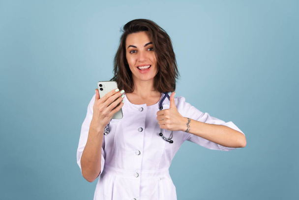 Νεαρή όμορφη γυναίκα γιατρός σε μια ρόμπα εργαστηρίου σε μπλε φόντο κρατώντας ένα τηλέφωνο, κοιτάζοντας την κάμερα, χαμογελώντας χαρούμενα - Φωτογραφία, εικόνα
