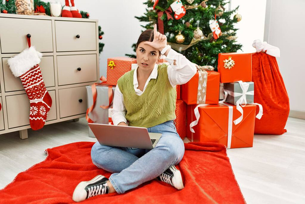Mladá krásná hispánská žena pomocí notebooku sedí u vánočního stromku dělat si legraci z lidí s prsty na čele dělá gesto poražený posměch a urážlivé.  - Fotografie, Obrázek