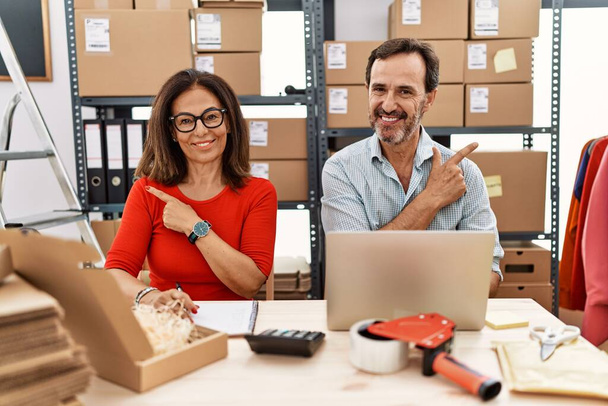 Middle age couple working at small business ecommerce cheerful with a smile on face pointing with hand and finger up to the side with happy and natural expression  - Photo, Image