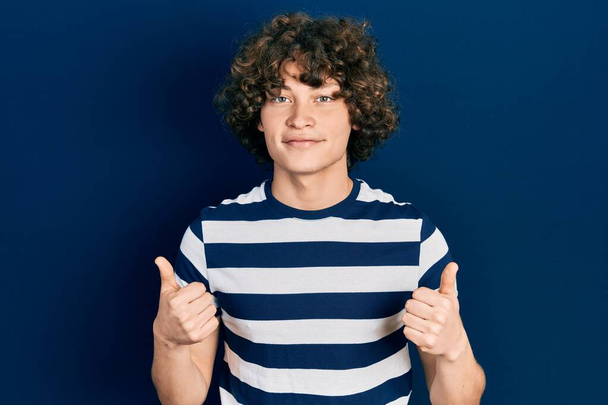 Handsome young man wearing casual striped t shirt success sign doing positive gesture with hand, thumbs up smiling and happy. cheerful expression and winner gesture.  - Photo, Image