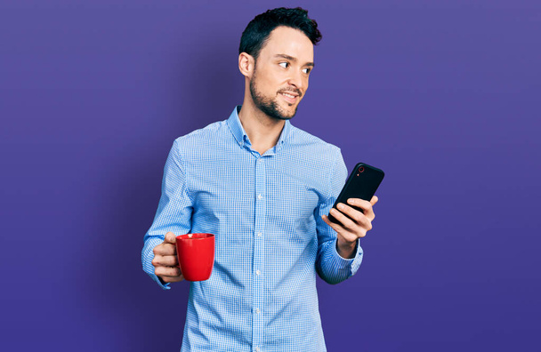 Hombre hispano con barba usando smartphone y bebiendo una taza de café sonriendo mirando a un lado y mirando a otro pensando.  - Foto, imagen