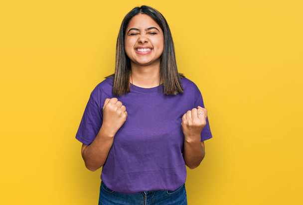 Jeune fille hispanique portant t-shirt violet occasionnel excité pour le succès avec les bras levés et les yeux fermés célébrant la victoire en souriant. concept gagnant.  - Photo, image