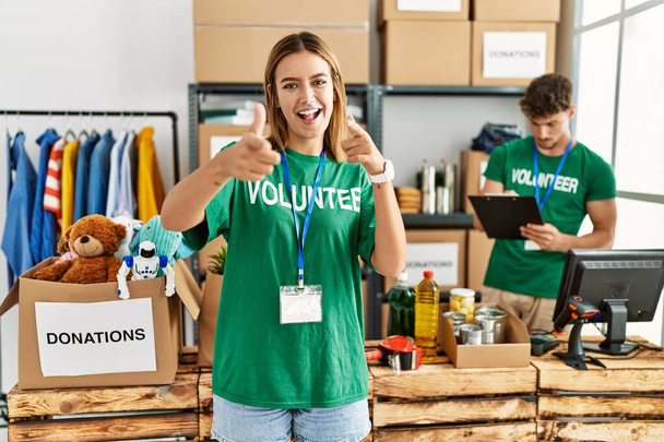 Young blonde girl wearing volunteer t shirt at donation stand pointing fingers to camera with happy and funny face. good energy and vibes.  - Photo, Image