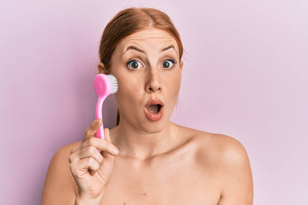 Young irish woman using facial exfoliating brush scared and amazed with open mouth for surprise, disbelief face  - Фото, изображение