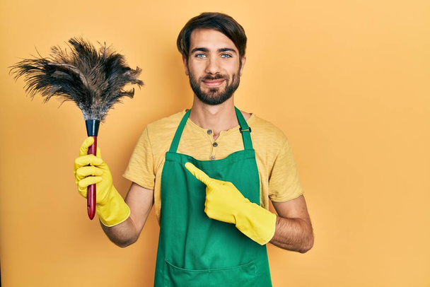 Önlük giyen genç İspanyol adam temizleme fırçası tutuyor. Gülümsüyor, el ve parmağıyla işaret ediyor.  - Fotoğraf, Görsel