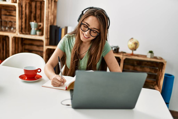 Young hispanic woman using laptop and writing on book sitting on table at home - 写真・画像