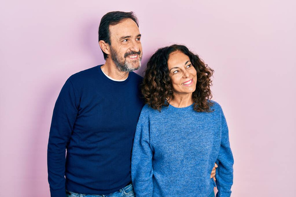Middle age couple of hispanic woman and man hugging and standing together looking away to side with smile on face, natural expression. laughing confident.  - Foto, Imagen