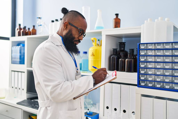 Junger afrikanisch-amerikanischer Mann trägt Wissenschaftleruniform mit Schriftzug auf Klemmbrett im Labor - Foto, Bild