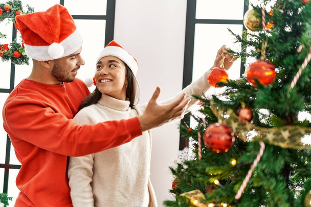 Joven pareja hispana sonriendo feliz decorando el árbol de Navidad en casa. - Foto, imagen