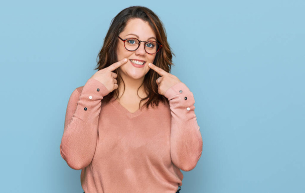 Young plus size woman wearing casual clothes and glasses smiling with open mouth, fingers pointing and forcing cheerful smile  - Photo, Image