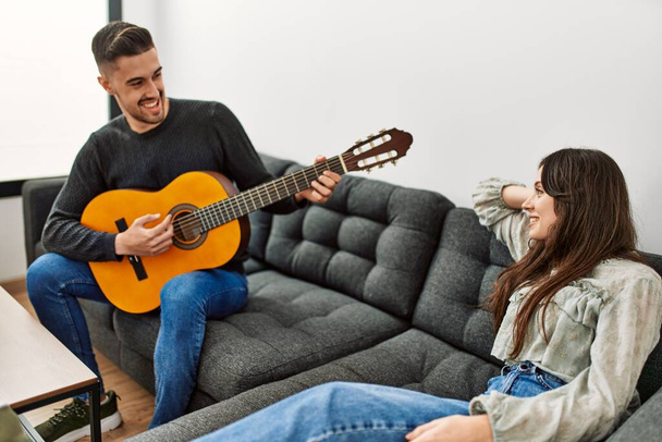 Mladý hispánský pár hraje španělskou kytaru sedí na pohovce doma. - Fotografie, Obrázek