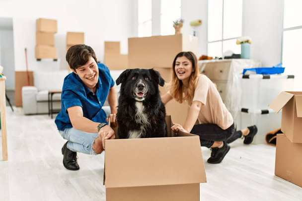 Joven pareja caucásica sonriendo feliz transportando perro en la caja de cartón como un coche en el nuevo hogar. - Foto, imagen
