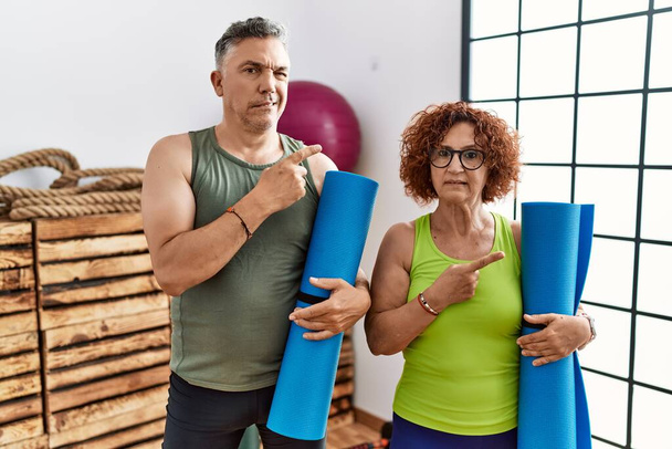 Middle age couple holding yoga mat pointing aside worried and nervous with forefinger, concerned and surprised expression  - Photo, Image