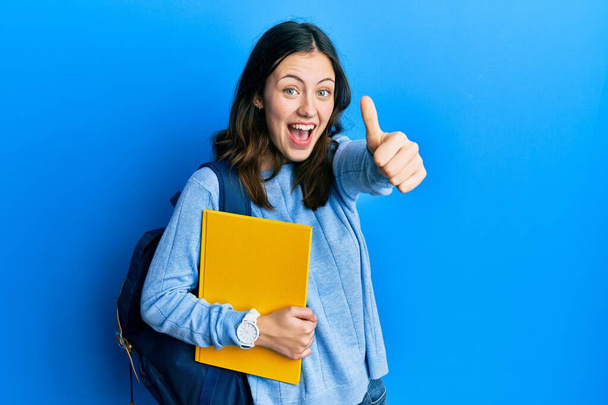 Young brunette student woman wearing student backpack doing thumbs up gesture celebrating crazy and amazed for success with open eyes screaming excited.  - Photo, Image