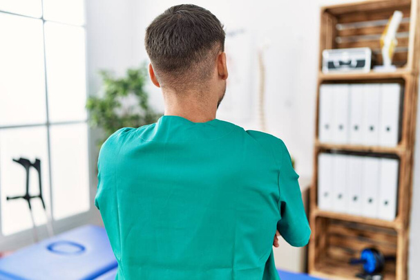 Młody fizjoterapeuta pracujący w klinice rekonwalescencji bólu stojący tyłem i patrzący z skrzyżowanymi ramionami  - Zdjęcie, obraz