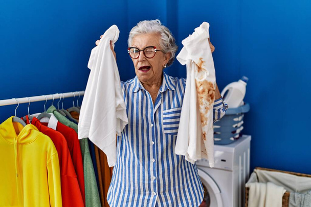 Mujer mayor con el pelo gris sosteniendo camiseta blanca limpia y camiseta con mancha sucia guiño mirando a la cámara con expresión sexy, cara alegre y feliz.  - Foto, imagen