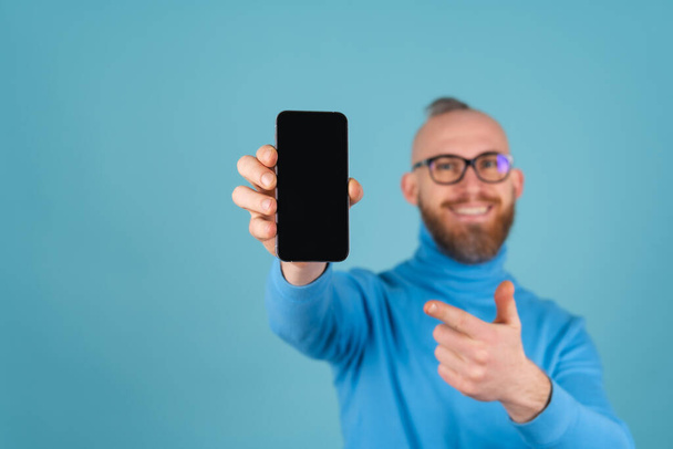 Un joven con una barba roja en un cuello alto sobre un fondo azul sostiene un teléfono con una pantalla negra en blanco para una inscripción en foco, se extiende a la cámara - Foto, Imagen