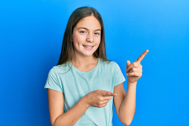 Belle petite fille brune portant un t-shirt blanc décontracté souriant et regardant la caméra pointant avec deux mains et doigts sur le côté.  - Photo, image