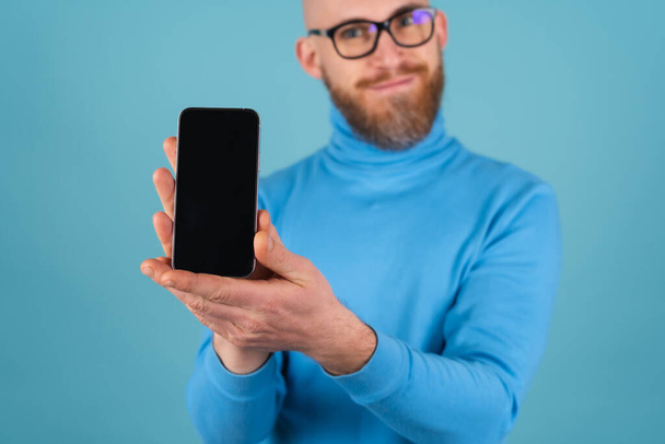 Mavi arka planda, boğazlı kazaklı, kırmızı sakallı genç bir adam odak noktasında bir yazı için boş siyah ekranlı bir telefon tutuyor ve kameraya uzanıyor. - Fotoğraf, Görsel