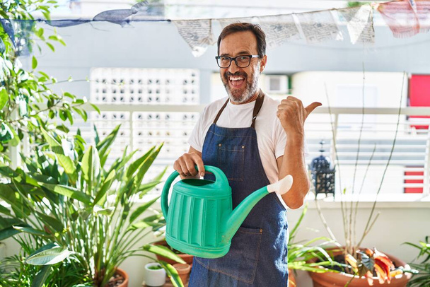 Hombre de mediana edad con delantal de jardinero sosteniendo regadera en casa terraza apuntando pulgar hacia el lado sonriendo feliz con la boca abierta  - Foto, imagen