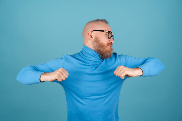 Un giovane uomo con la barba rossa con un dolcevita su uno sfondo blu sta ballando allegramente, divertendosi - Foto, immagini