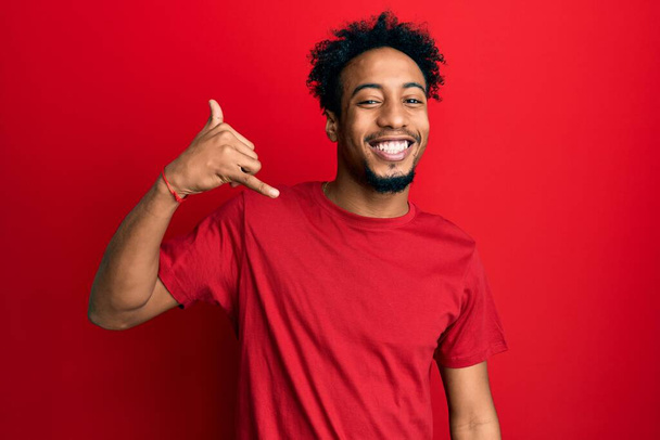Ein junger afrikanisch-amerikanischer Mann mit Bart trägt ein lässiges rotes T-Shirt, lächelt und macht Telefongesten mit Händen und Fingern, als würde er telefonieren. Vermittlung von Konzepten.  - Foto, Bild