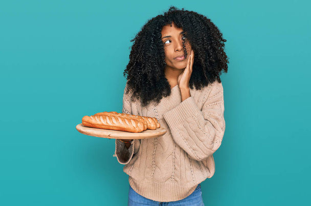 jong Afrikaans amerikaans meisje houden lade met brood serieus gezicht denken over vraag met de hand op kin, attent over verwarrend idee  - Foto, afbeelding