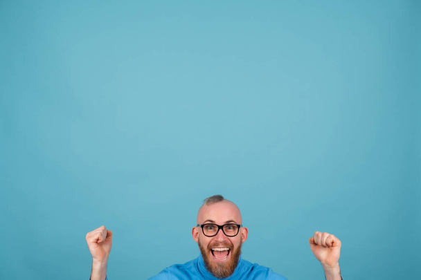 Egy vörös szakállú fiatalember egy kék hátterű garbóban vidám, sikoltozva a boldogságtól, egy győztes gesztusa. - Fotó, kép