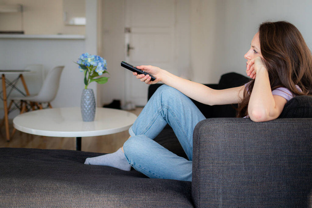 Скучающая женщина использует пульт дистанционного управления, чтобы смотреть телевизор дома. Человек не может решить, какой канал смотреть - Фото, изображение