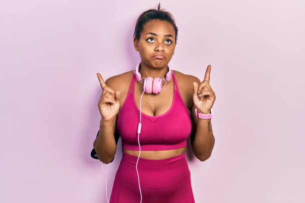 Young african american girl wearing gym clothes and using headphones pointing up looking sad and upset, indicating direction with fingers, unhappy and depressed.  - Photo, Image