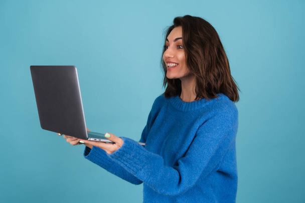Junge Frau im Strickpullover hält vereinzelt einen Laptop in der Hand, blickt überrascht auf den Bildschirm - Foto, Bild
