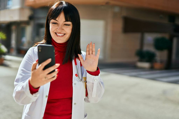 Mladá brunetka žena nosí lékařskou uniformu dělá videokall s smartphonem ve městě - Fotografie, Obrázek