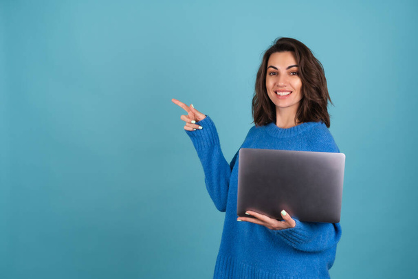 Młoda kobieta w swetrze z dzianiny trzyma laptopa, patrzy na ekran i uśmiecha się radośnie, wskazując puste miejsce po lewej stronie - Zdjęcie, obraz