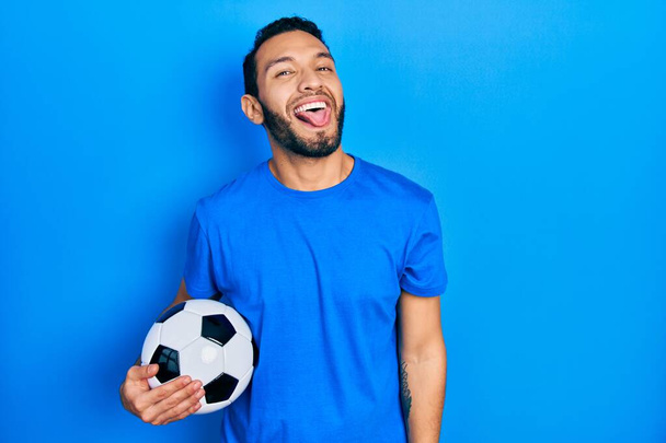 Ισπανόφωνος με μούσι που κρατάει μπάλα ποδοσφαίρου και βγάζει γλώσσα χαρούμενος με αστεία έκφραση. έννοια συναισθήματος.  - Φωτογραφία, εικόνα